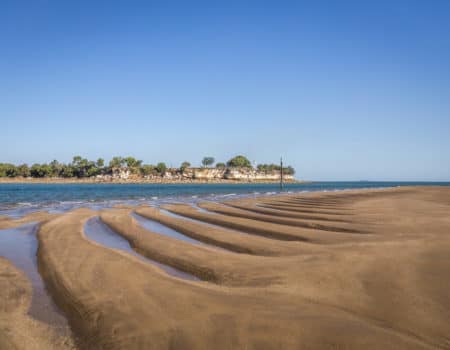 Curved tidal sand ripples along the beach in Darwin with blue sky, calm water and a distant rocky headland