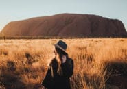 Female traveller in wide-brimmed hat gazing at Uluru at sunrise, surrounded by golden spinifex grass
