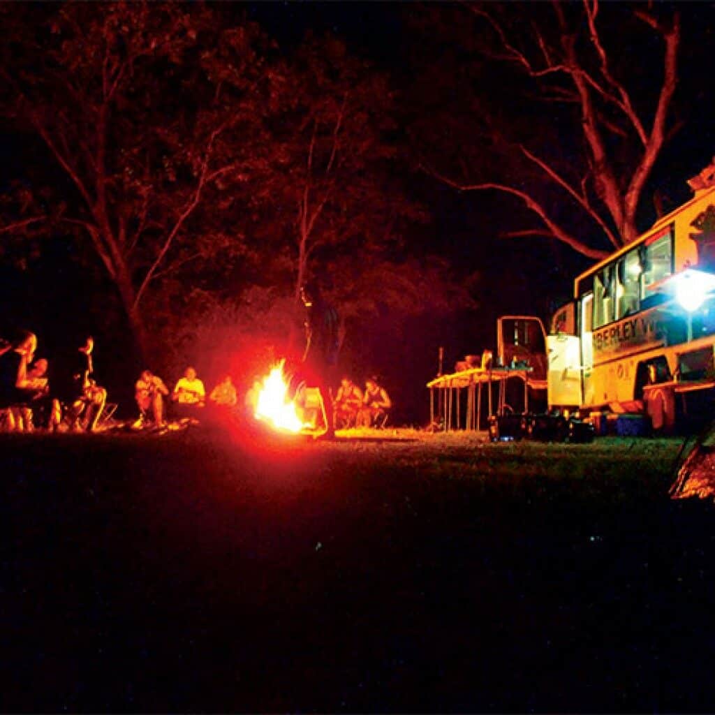 Group of travellers gathered around a glowing campfire at night beside a Kimberley Tours overland bus under the trees in outback Australia.